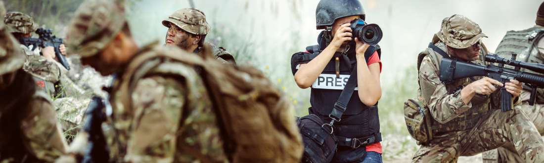 Image shows an embedded combat journalist wearing a bullet proof vest and taking photos, surrounded by special forces with guns