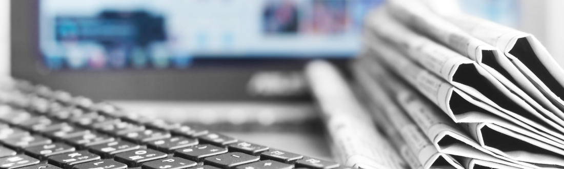 Image shows a stack of newspapers and a keyboard with an out of focus computer screen in the background